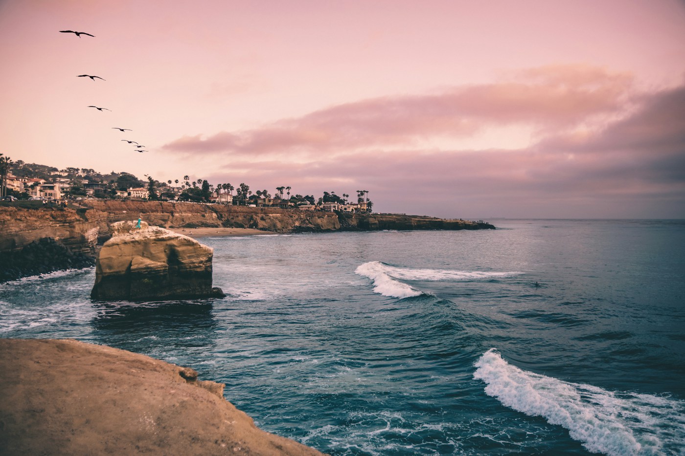 surfers waiting beside breaking waves at sunset cliffs beneath a pink evening sky